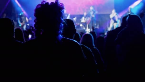 Multidão de pessoas ficam perto de um palco aberto olhando um concerto de rock à noite — Vídeo de Stock