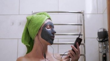 Yüzünde kil maskesi olan bir kız Smartphone 'da kulaklıkla müzik dinliyor.
