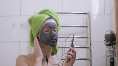 Yüzünde kil maskesi olan bir kız Smartphone 'da kulaklıkla müzik dinliyor.