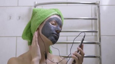 Yüzünde kil maskesi olan bir kız Smartphone 'da kulaklıkla müzik dinliyor.