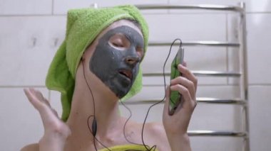 Yüzünde kil maskesi olan bir kız Smartphone 'da kulaklıkla müzik dinliyor.
