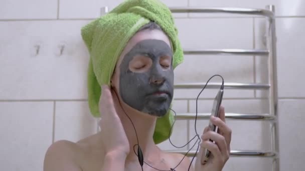 Dziewczyna z maską Clay on Face Słucha muzyki ze słuchawkami na smartfonie — Wideo stockowe