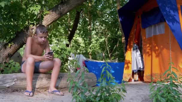 Niño triste se sienta en un tronco, jugando en un teléfono inteligente en un bosque en el sol cerca de la tienda — Vídeos de Stock