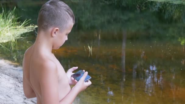 슬픈 아이는 강둑에 머물면서 숲 속의 스마트 폰으로 놀고 있다. 줌 — 비디오