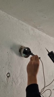 Macro vertical shot of an electricians hand testing voltage at an electrical outlet box on a white textured wall, showing red indicator lights flashing on the tester.