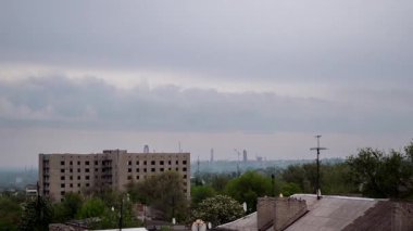 Timelapse shot over old city rooftops towards an unfinished concrete structure, showing empty window sockets, distant factory cranes, and fast-moving clouds across a grim, desaturated grey sky.