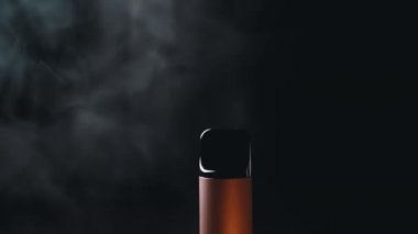 Yakın plan, Elektronik Sigara veya Siyah Arkaplanda Tüttüren Vape. Karanlık bir ortamda ortaya çıkan dumanlı tek bir cisim gizemli bir atmosfer yaratıyor. Doku. Bulanık hareket.