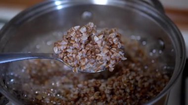 Ev mutfağında karabuğday lapası pişirme işlemi. Bir kadın elini karıştırır ve tahıldan yapılan lapayı kaşıkla kontrol eder.