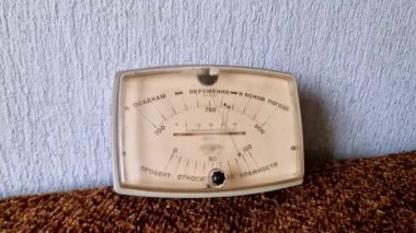 Kiril alfabesiyle yazılmış kahverengi desenli bir halının üzerinde duran klasik bir analog barometreye yakın plan. İğne açık mavi duvar arka planına karşı değişken hava koşullarını gösterir.