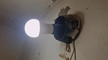 Parçalanmış beyaz alçı bir duvara monte edilmiş eski siyah bir armatürde aydınlanan modern LED ampulün yakın plan ayrıntıları. Lambanın altında gevşek iki elektrik kablosu asılı..