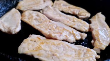 Yukardan yakından çekilmiş çiğ tavuk göğsü şeritleri siyah bir tavada yağda kızartılıyor. Kümes hayvanları cızırdar ve yemek hazırlıkları sırasında fırında yemek pişirirken yavaşça kahverengiye döner..