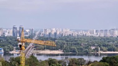 Geniş bir nehir ve köprüye bakan sarı bir kafes inşaat vincinin yüksek açılı görüntüsü. Arka planda, yüksek katlı apartmanların yoğun bir şehir silueti ufuk boyunca uzanır..