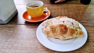 Beyaz bir tabakta rendelenmiş peynirle kaplı lezzetli bir kruvasanın statik çekimi. Yanındaki sarı kahve, ahşap bir kafe masasında, kahvaltı ya da öğle yemeği molası için hazır..