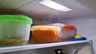 Soğuk buharın ya da plastik konteynırlardan arta kalan yiyeceklerle birlikte buharın yükseldiği bir buzdolabının içinden bak. Aydınlatılmış rafta bir kap doğranmış havuç ve bir limon görülebilir..