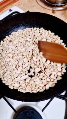 Dikey, yüksek açılı bir tahta spatula görüntüsü sıcak siyah dökme demir tavada çiğ fıstık karıştırıyor. Sosyal medya içeriği için mutfaktaki gaz sobasında fındık kızartma işlemi..