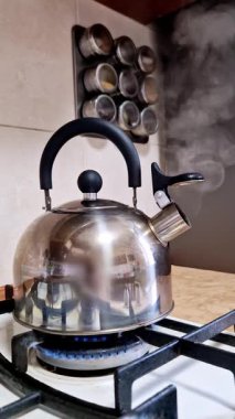 Paslanmaz çelik çaydanlık gaz sobasında kaynıyor ve tencerenin altında parlak mavi bir alev görünüyor. Sıcak bir içecek için modern bir mutfak ortamında su ısındıkça, su borusundan sürekli buhar yükseliyor..