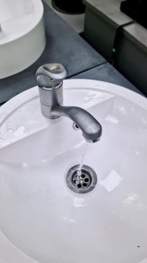 Temiz, beyaz seramik bir lavaboya sürekli akan, yıpranmış krom bir musluğun yüksek açılı görüntüsü. Su, modern bir banyoda ya da umumi tuvalette doğrudan metal kanalından akıyor..