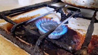 Siyah ızgaralı ve lekeli yüzeyi olan kirli beyaz bir sobanın üzerinde yanan mavi bir gaz alevinin yakın çekimi. Bu mutfak aletleri ev enerjisi tüketimini ve kamu maliyetlerini temsil ediyor..