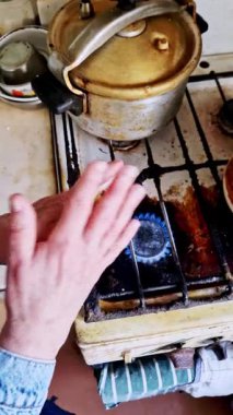 Mavi bir gaz alevi üzerinde ısınan yaşlı ellerin dikey ve tepeden görünüşü. Kot ceket manşeti görünür. Kirli beyaz gaz ocağının arka planında ısıtma konseptini temsil eden bir düdüklü tencere var..