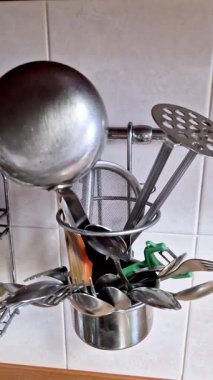 Paslanmaz çelik bir tutacağa yerleştirilmiş çeşitli mutfak aletlerinin dikey ve yakın çekim görüntüleri. Bir kepçe, patates ezici, süzgeç, kaşıklar ve çatallar fayanslı bir mutfak duvarına karşı demiryolu üzerinde saklanır..