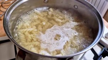 Sarı fusilli makarnanın yukarıdan görünüşü paslanmaz çelik bir tencerede güçlü bir şekilde kaynıyor. Temiz su bol kabarcıklar ve beyaz nişasta köpüğü ile çalkalanır. Bu da aktif bir mutfak hazırlığını gösterir..