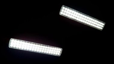 İki dikdörtgen LED ışık çubuğu, parlak diyotlardan oluşan bir ızgarayı ortaya çıkararak, koyu siyah bir arkaplana karşı aydınlanıyor. Beyaz ışığın hafif bir titreşimi var, modern, teknolojik bir his yaratıyor..