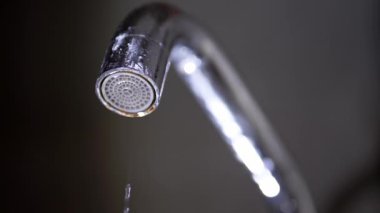 Krom su musluğu sızdıran bir musluktan bir damla su damlatır. Makro çekim, ayrıntıları koyu ve bulanık bir arkaplanla yakalar.