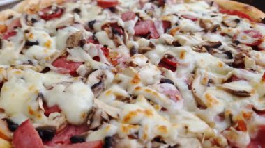 Taze pişmiş pizza eritilmiş mozarella peyniri, sosis ve mantarla kaplıdır. Kamera yavaşça yaklaşır, malzemelerin iştah açıcı dokularına odaklanır..