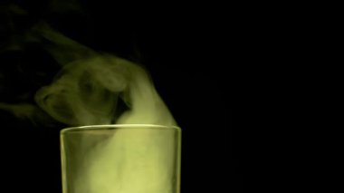 Kalın sarı duman, şeffaf bir cam şişeden koyu bir arka plana doğru yavaşça yükselir. Yan görüş. Tehlikeli kimyasal reaksiyonlar, zehirli deneyler ve gizemli sihirli iksir kavramı.