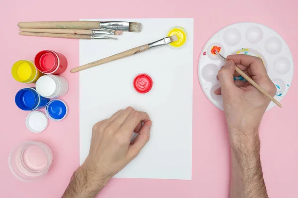 Erkek el, akrilik boyalı beyaz bir kağıt üzerinde fırçayla resim çizer. Palette suluboya karıştırıyorum. Pembe arkaplan.