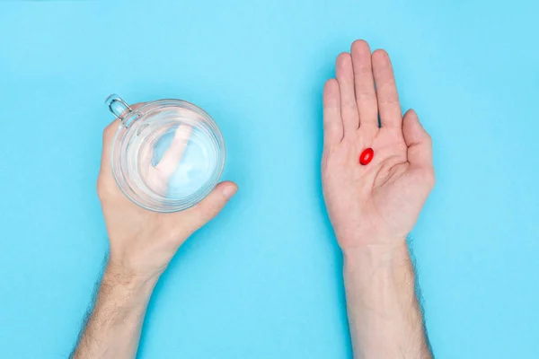 Omul ţine o pastilă roşie şi un pahar cu apă. Fundal albastru. Contextul medical. Luând medicamente . — Fotografie, imagine de stoc