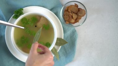 A womans hand adds bay leaf to a bowl of broth. Parsley leaves and croutons.