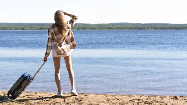 Young woman with her suitcase luggage on the sandy shore of a lake or river on sunny day looks into the distance. Hipster female in loose cotton shirt and shorts is ready to travel. Journey concept — Stockfoto