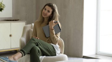 Dijital tablet kullanan, koltukta oturan, gülümseyen ve bilgisayar tutan genç bir kadın..