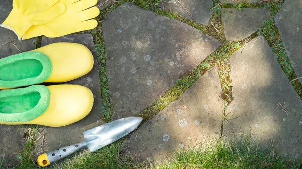 Equipo de jardinería. Pala o pala de jardín, guantes de mano amarillos y zapatos de botas de goma en el camino de piedra en el jardín. Fondo de granja. Vista superior. Copiar espacio. Enfoque selectivo. — Foto de Stock