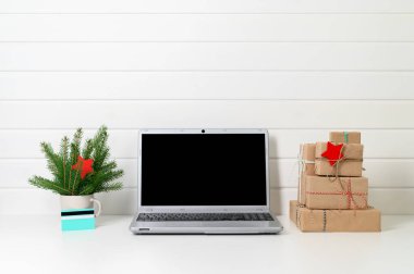 Laptop, hediyeler, kredi kartı ve ladin dalları beyaz ahşap bir duvarın arkasında..