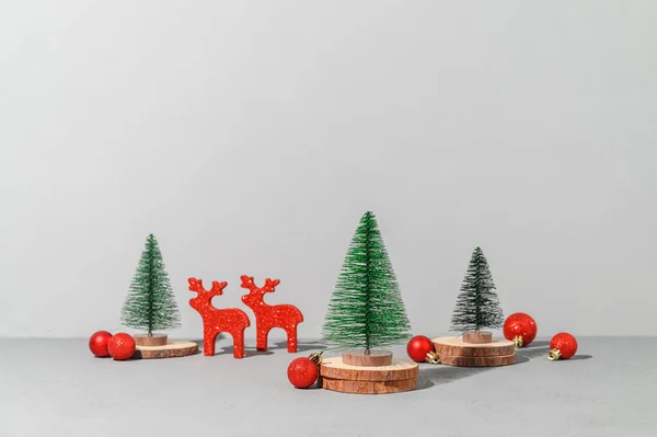 Conceptual festive composition with miniature Christmas trees on wooden podiums and red xmas decorations. Gray background. Copy space, place for text