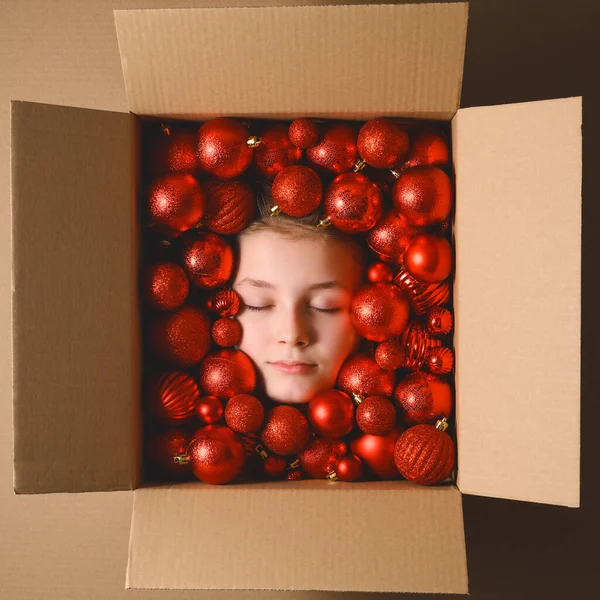 Creative Christmas composition. Sleeping head of teenage girl in cardboard box with red Christmas balls. Top view, vertical orientation