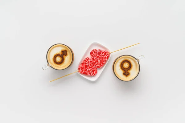 Two cups of coffee with symbols of venus and mars on whipped milk foam and couple lollipops in heart shape on white tabletop. Concept romantic sweet breakfast on Valentine\'s day. Flat lay, copy space
