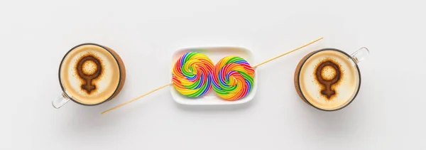 Two cups of coffee with symbols of venus on whipped milk foam and couple round rainbow lollipops on white tabletop. Concept lesbian love. Flat lay, top view, banner. LGBT