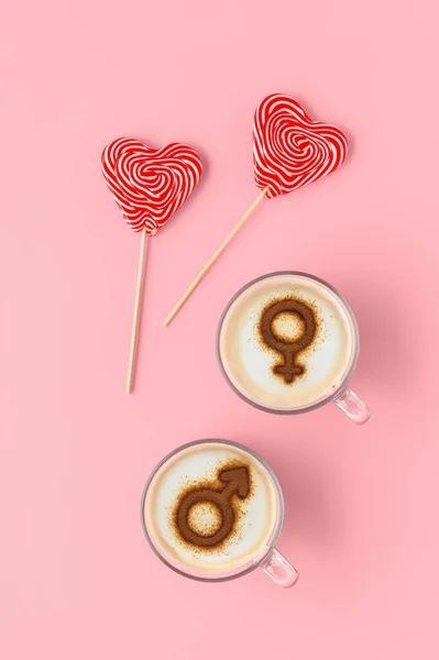 Two cups of coffee with symbols of venus and mars on whipped milk foam and couple of lollipops in heart shape. Pastel pink table. Concept romantic sweet breakfast on Valentine\'s day. Flat lay