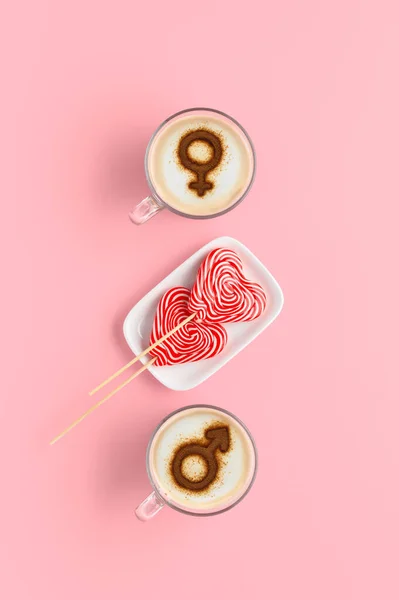 Two cups of coffee with symbols of venus and mars on whipped milk foam and couple of lollipops in heart shape. Pastel pink background. Concept romantic breakfast on Valentine\'s day. Flat lay