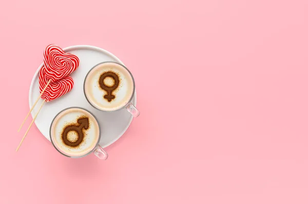 Two cups of coffee with symbols of venus and mars on whipped milk foam and couple of lollipops in heart shape. Pastel pink background. Concept romantic breakfast on Valentine\'s day. copy space
