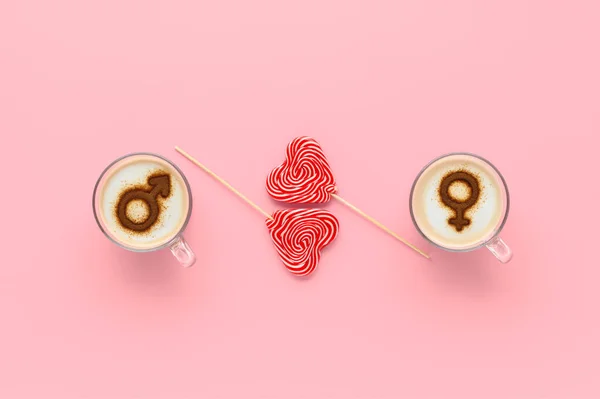 Two cups of coffee with symbols of venus and mars on whipped milk foam and couple of lollipops in heart shape. Pastel pink background. Concept romantic breakfast on Valentine\'s day. Flat lay