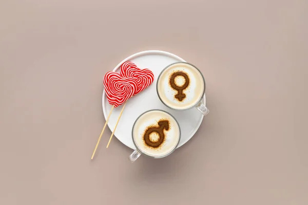 Two cups of coffee with symbols of mars and venus on whipped milk foam and lollipops in heart shape on white plate. Beige background. Concept romantic date on Valentine's day. Flat lay