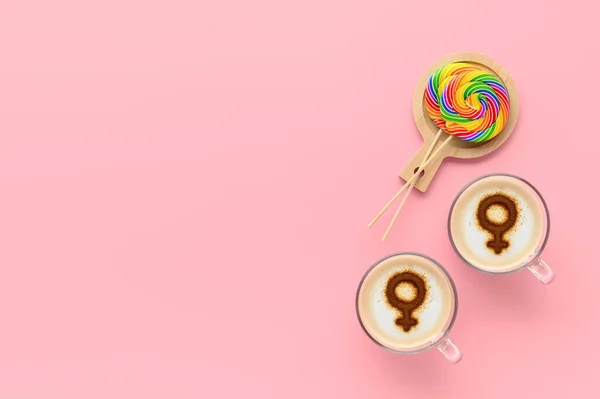 Two cups of coffee with symbols of venus on whipped milk foam and couple round LGBT rainbow lollipops on pastel pink background. Concept lesbian love. Flat lay, top view, copy space