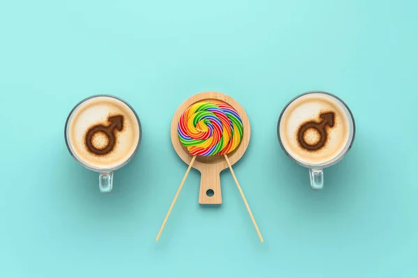 Two cups of coffee with symbols of mars on whipped milk foam and couple round LGBT rainbow lollipops on pastel cyan background. Concept gay love. Flat lay, top view