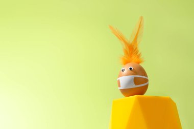 Yaratıcı Paskalya konsepti. Yumurtadan yapılan Paskalya tavşanı yeşil arka planda koruyucu yüz maskesi takıyor. Yumurta sarı bir kaidede dengelenmiş. Boşluğu kopyala