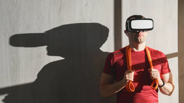 Fitness, workout and VR technology. Portrait Caucasian young adult man in a red t-shirt with an expander on the shoulders, wearing virtual reality glasses. Indoors, copy space