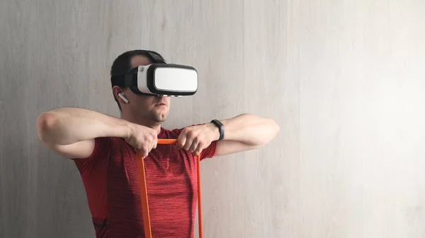 Fitness, workout and vr technology. Caucasian young adult man in a red t-shirt training in home, using expander, wearing virtual reality glasses. Indoors